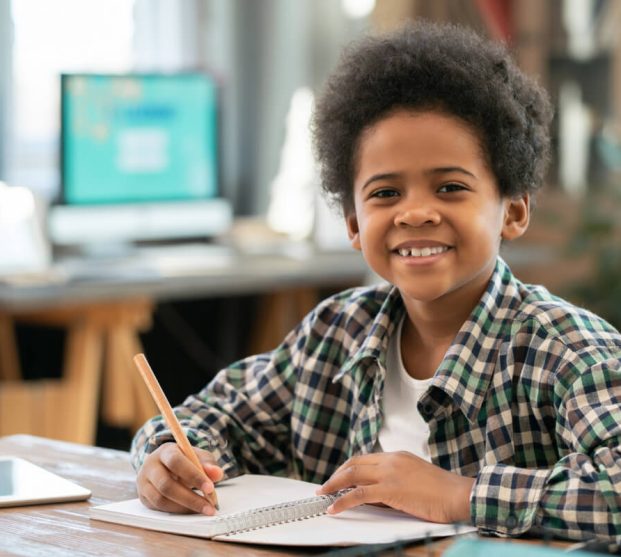 adorable-schoolboy-african-ethnicity-making-notes-copybook-looking-you-with-smile-while-doing-homework-by-table-living-room