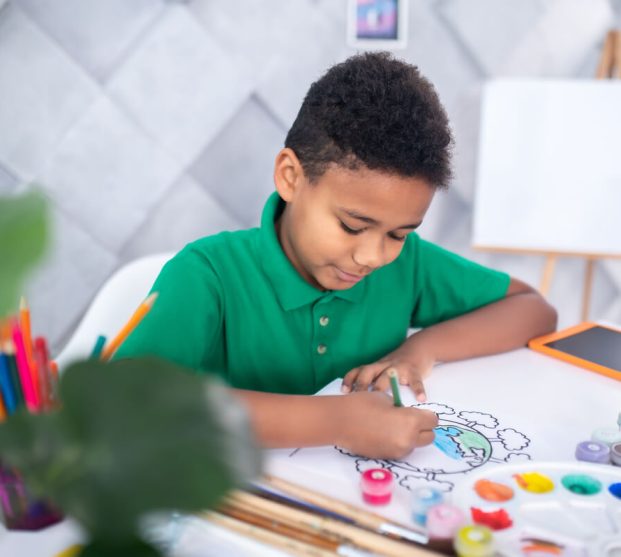 enthusiasm-dark-skinned-curly-haired-boy-green-tshirt-enthusiastically-sketching-picture-with-colored-pencil-sitting-table-light-room